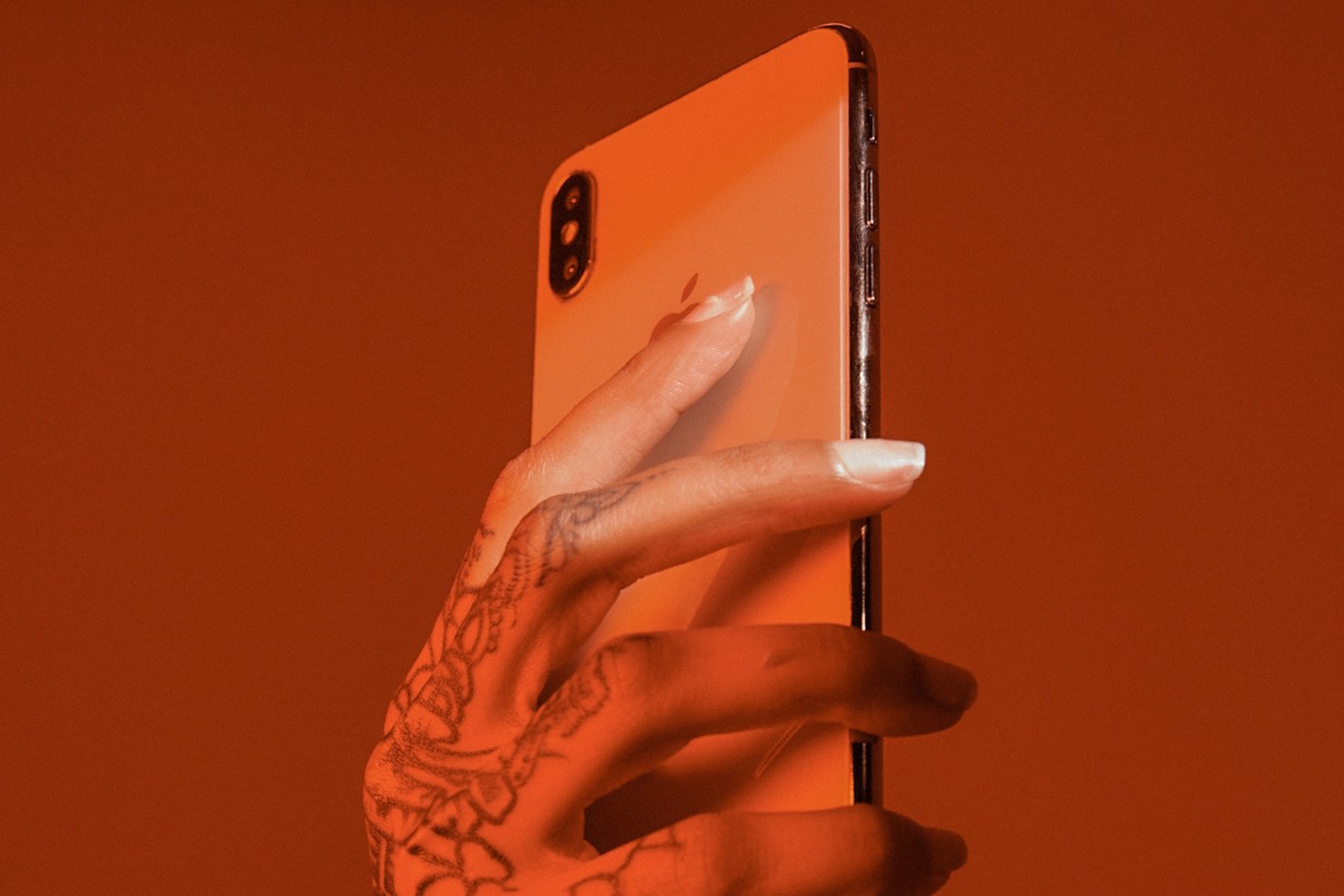 A rusty red photograph of a hand with an artistic henna tattoo holding an iPhone in the air, with a beam of white light illuminating the index and middle finger.